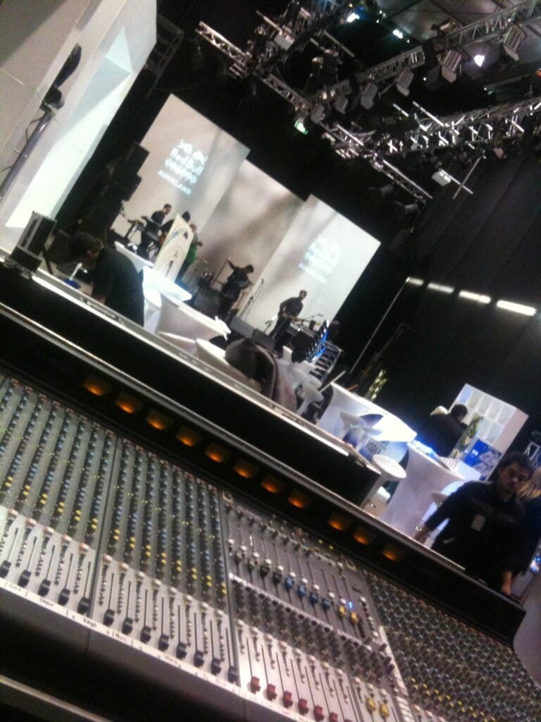 A large mixing desk in the foreground, and a stage is being set up with a sound system and screens in the background.