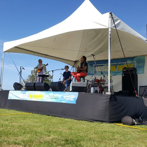 Hire a Stage, marquee, and soundsystem, with a band playing at an outdoor event. An example of stage hire Auckland. The Rock Factory's stage hire services are perfect for any event. This photo shows the 6 by 6 springtop marquee hire auckland option placed on top of a 6 by 6 stage.