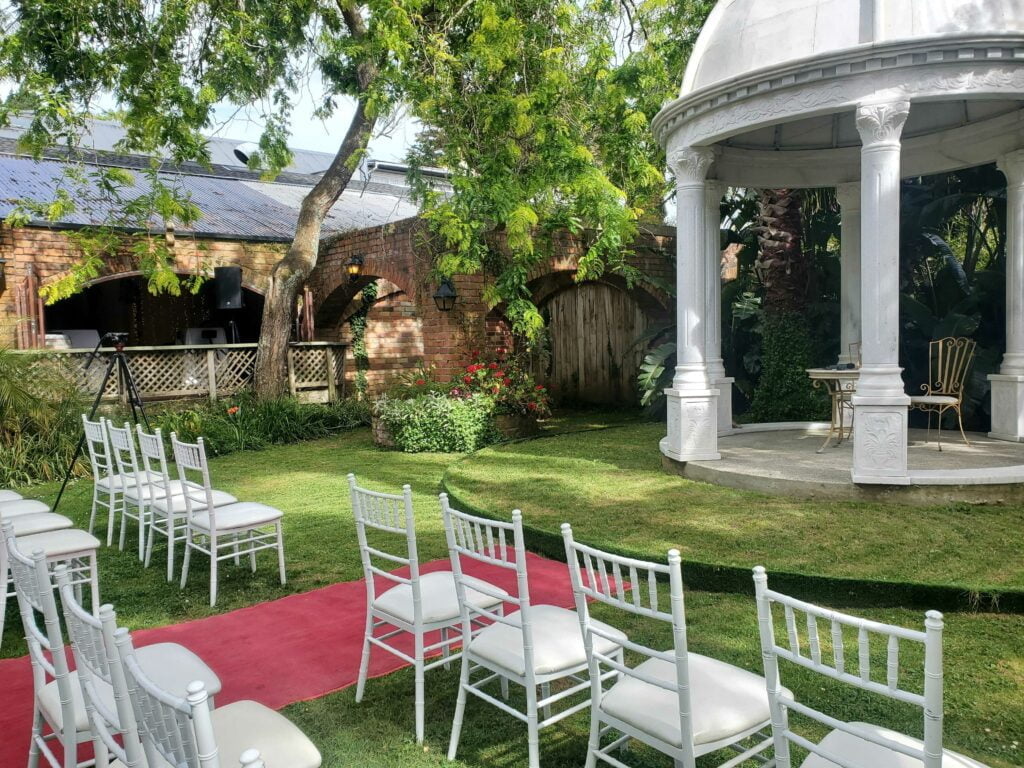 Outdoor wedding set-up, beautiful chairs and domed pagodoa are prominent, and microphones, speakers, and a camera can be seen subtly in the background.