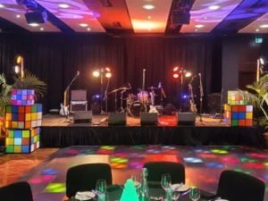 80's themed corporate party event at the end-of-year celebration. Colourful Rubix cube decorations are around the stage, and lighting matches these. There is a stage with a drumkit set up for a band. Business event end of year function or expo example. Hire entertainment and artist booking agent services available.