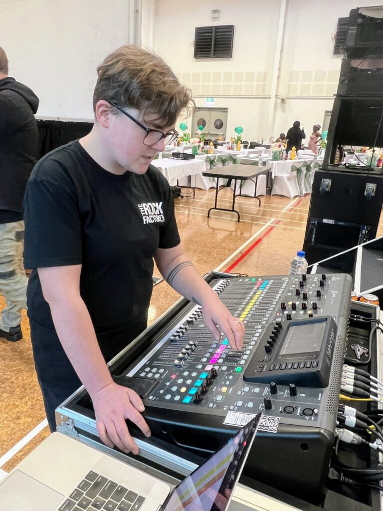 One of our technicians PJ behind the mixing desk used at hall hire auckland. The photo shows an X32 sound desk, laptop for playback, and you can see the back of one of the speaker stacks for one of our concert PA sound systems.